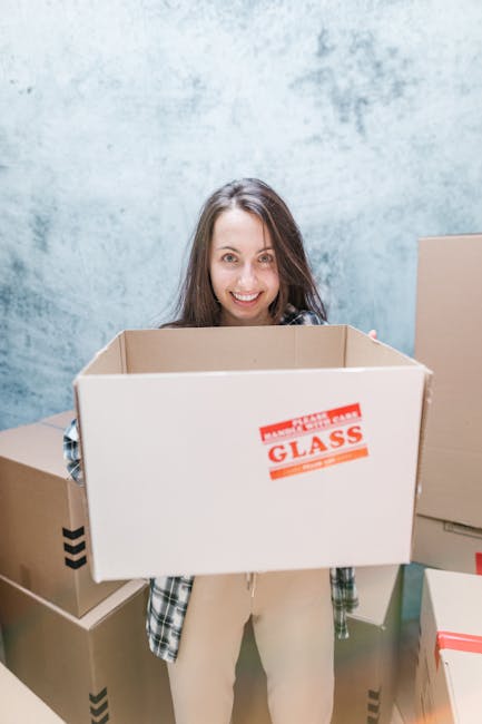 A smiling woman with long brown hair, dressed in casual clothing, is standing indoors in front of a light blue textured wall. She is holding an open cardboard box in front of her, which has a red and white label reading 'FRAGILE GLASS' attached to one side. Surrounding her are several other closed cardboard boxes of varying sizes, some sealed with packing tape and others partially open, indicating a home relocation or packing scenario. The environment appears well-lit, capturing the busy process of packing for a house move, with the boxes positioned on the floor and some leaned against the wall. This scene exemplifies the careful preparation involved in furniture transport and the packing process typical of professional removals services provided by Man and Van Holland Park. The focus on the woman and the boxes aligns with themes of packing and moving logistics necessary for house removals in Holland Park.