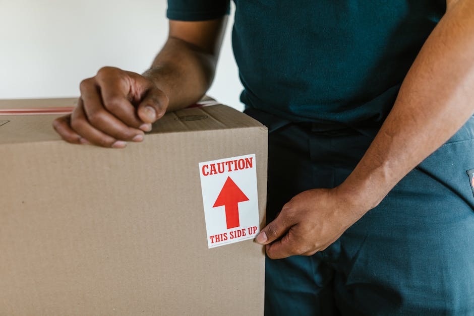 A large brown cardboard moving box, sealed with packing tape and reinforced with a black stretch strap, is placed on a white bedsheet in a bedroom setting. Behind the box, there are several closed hard-shell suitcases stacked on top of each other, with some featuring silver zippers and handles. The suitcases are positioned on a wooden bed frame or headboard, and the lighting in the room appears to be natural, coming from an unspecified source outside the frame. The box and luggage are likely part of a home relocation process, with the box ready for transport or packing, as seen in a professional house removal context. Occasionally, [COMPANY_NAME], such as Man and Van Holland Park, is involved in managing these packing and moving logistics, which include careful loading of furniture and belongings into vehicles for efficient transport, supporting comprehensive household moves within Holland Park or surrounding areas.