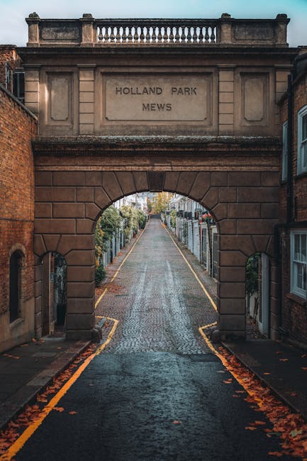 A cobbled lane leading through a stone archway inscribed with 'Holland Park Mews,' flanked by residential buildings and private gardens. The street appears slightly wet, with fallen autumn leaves scattered along the pavement. In the foreground, a moving trolley loaded with cardboard boxes and wrapped furniture is partially visible, indicating a home relocation process. The scene is lit by natural daylight, with soft shadows cast on the street, capturing a typical setting for furniture transport and packing during a house move. The image emphasizes the narrow, quiet street typical of Holland Park Mews, aligning with professional removals services offered by Man and Van Holland Park, who handle loading and transportation within such environments.