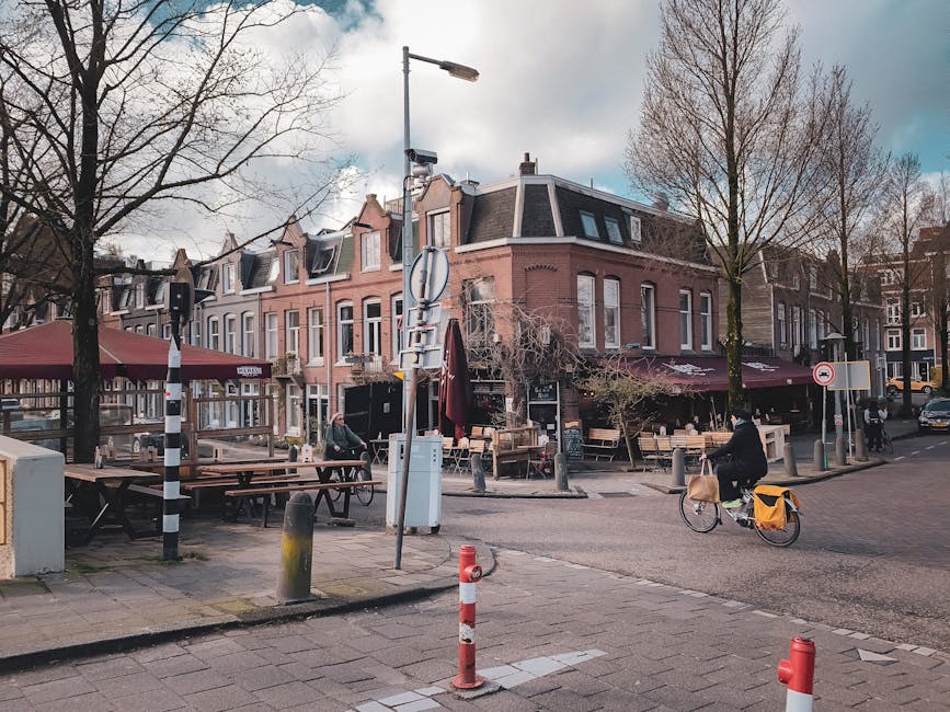 A street scene in Holland Park showing a row of three-storey residential buildings with classic brick facades, sloped roofs with dormer windows, and parked bicycles alongside. The foreground features a sidewalk with outdoor seating areas, including wooden benches and tables under large maroon umbrellas, designated for a cafe or restaurant. A woman riding a bicycle with a yellow pannier basket is visible on the street, while trees with bare branches line the pavement, indicating a season outside of summer. Various traffic devices, such as black and white striped bollards, a street lamp, and a traffic camera, are positioned along the road and pavement. The area appears quiet with minimal pedestrian and vehicle movement, and the scene is captured during an overcast day with muted natural lighting. This setting exemplifies typical urban surroundings applicable to home relocation and furniture transport, where careful planning may be needed during moving logistics, as managed by Man and Van Holland Park, to avoid hidden costs associated with city-based removals.