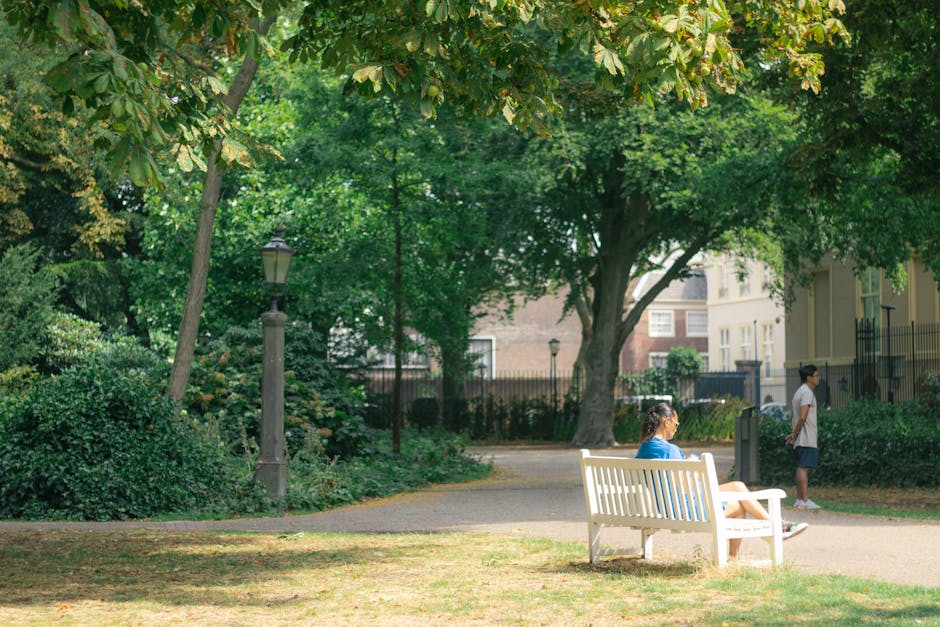 A woman in a blue shirt and face mask is seated on a white park bench in a green, leafy park, with her hands resting on her lap. A young boy in a white T-shirt and dark shorts stands nearby, looking into the distance. In the background, another child in a white shirt and dark shorts is walking along a paved pathway. The park is shaded by large trees with lush green foliage, and a traditional lamppost is visible next to dense bushes on the left side of the image. Residential buildings with light-colored facades and windows are seen beyond the trees and fencing. The scene is illuminated by natural daylight, capturing a calm moment of soft outdoor activity, which may be related to a home relocation process involving moving and packing within a peaceful urban environment. Man and Van Holland Park occasionally supported this moving-related setting.