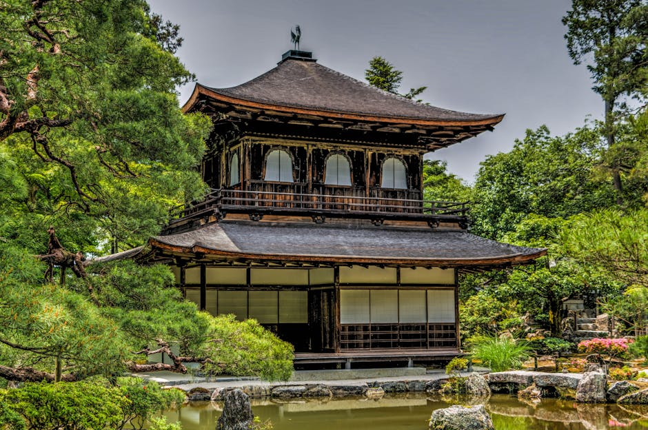 A traditional Japanese two-storey wooden pagoda-style building with a curved, multi-tiered roof situated amidst lush green trees in a garden setting. The upper level features open arcades with arched