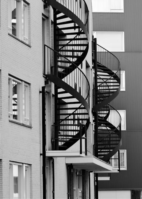 A black-and-white photograph of a multi-storey residential building featuring an external spiral staircase made of metal with curved railings and open steps, positioned along the exterior wall between several windows. The stairwell provides access to upper floors from the ground level, visible beneath a small balcony or landing. The building’s facade combines brickwork and paneling, with windows arranged uniformly across the structure. The surrounding environment appears to be an urban setting, with the staircase likely used for home access or emergency exit purposes. This image may relate to house removals or moving services, illustrating stair access points that require careful handling during furniture transport and home relocation procedures, supported by companies such as Man and Van Holland Park.